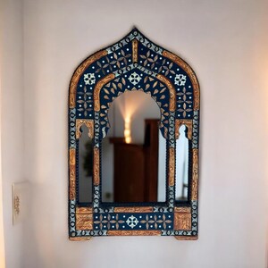 May include: An arched wall mirror featuring a dark blue frame with intricate gold and silver-toned floral and geometric carvings. The frame has a pointed top and decorative side cutouts, surrounding a central reflective glass surface.
