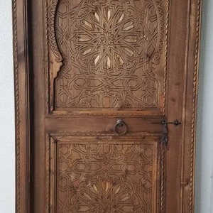 May include: A wooden door with intricate carved designs in a traditional Moroccan style. The door is made of dark wood and has a carved archway with a geometric pattern.