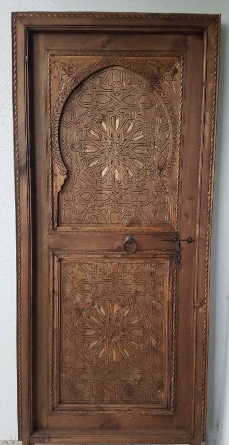 May include: A wooden door with intricate carved designs in a traditional Moroccan style. The door is made of dark wood and has a carved archway with a geometric pattern.