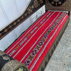 May include: A red and black patterned couch with ornate metal detailing and a wooden frame. The couch has a high back and a curved armrest. The metal detailing is silver and gold and features geometric designs.