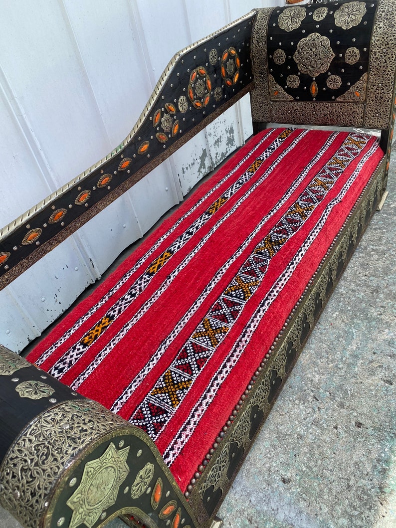 May include: A red and black patterned couch with ornate metal detailing and a wooden frame. The couch has a high back and a curved armrest. The metal detailing is silver and gold and features geometric designs.