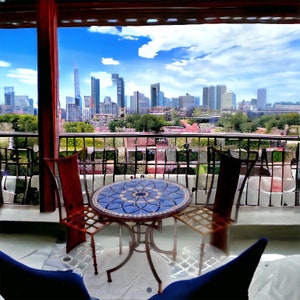 May include: A balcony scene featuring a small round table with a blue mosaic top and two matching chairs. The chairs have a reddish-brown metal frame. The background shows a cityscape under a blue sky.