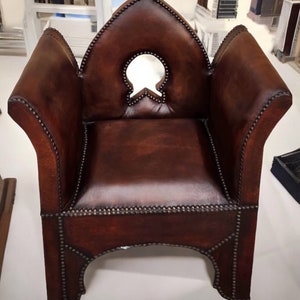 May include: A brown leather armchair with a pointed back and a decorative cutout in the back. The chair has a studded trim and is sitting on a white surface.