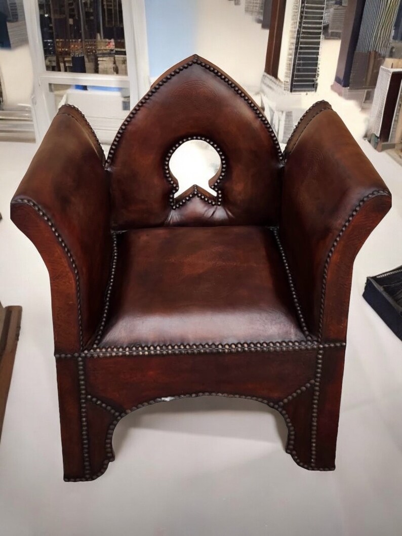 May include: A brown leather armchair with a pointed back and a decorative cutout in the back. The chair has a studded trim and is sitting on a white surface.