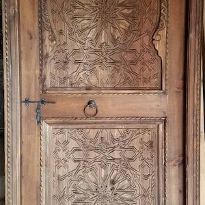 May include: A wooden door with intricate carved designs in a traditional Moroccan style. The door is brown and has a metal handle.