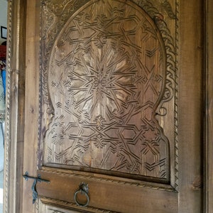 May include: A close-up of a wooden door with intricate carvings in a geometric pattern. The door is brown and has a metal handle.