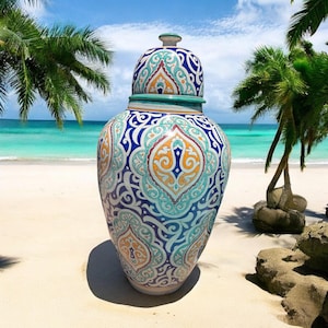 May include: A large, ornate ceramic vase with a lid, featuring a detailed pattern in blue, teal, orange, and white. The vase is set on a sandy beach with palm trees and a turquoise ocean in the background.