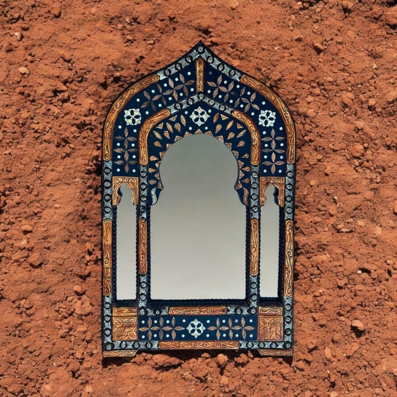 May include: An arched wall mirror featuring a dark blue frame with intricate gold-toned floral and geometric patterns. The frame includes white inlaid accents and a pointed top, surrounding a central reflective glass surface.