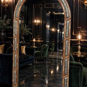 May include: A tall, arched floor mirror featuring a wooden frame with intricate, silver-toned metal inlay panels. The mirror has a distinct, curved top design and a wide, sturdy base, reflecting a dark, opulent room with velvet furniture.