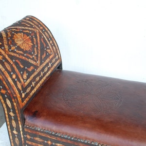 May include: A brown leather bench with a carved wooden back and seat. The back is decorated with a geometric pattern and the seat has a circular design.