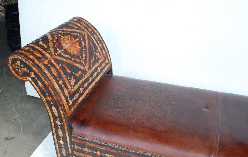 May include: A brown leather bench with a carved wooden back and seat. The back is decorated with a geometric pattern and the seat has a circular design.