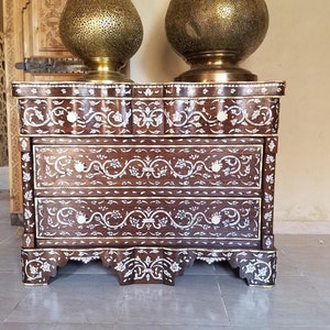 May include: A three-drawer chest with intricate floral designs inlaid with mother-of-pearl. The chest is made of dark wood and has a scalloped base.