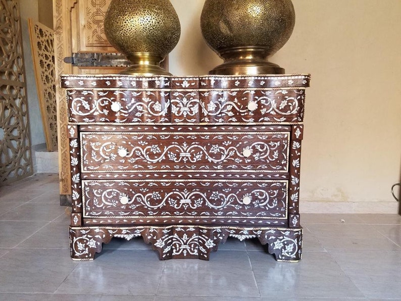 May include: A three-drawer chest with intricate floral designs inlaid with mother-of-pearl. The chest is made of dark wood and has a scalloped base.