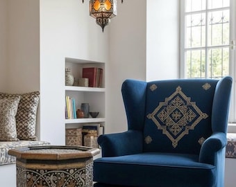 May include: A cozy living room scene featuring a navy blue wingback chair with gold embroidery, a decorative side table, and a hanging chandelier with ornate, amber-toned lanterns. The room has a neutral color palette with a patterned rug.