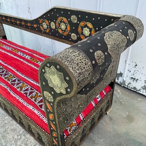 May include: A black and silver ornate couch with a red and white patterned cushion. The couch has intricate metalwork and is decorated with small, round, orange and silver accents.