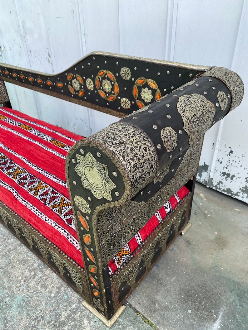 May include: A black and silver ornate couch with a red and white patterned cushion. The couch has intricate metalwork and is decorated with small, round, orange and silver accents.