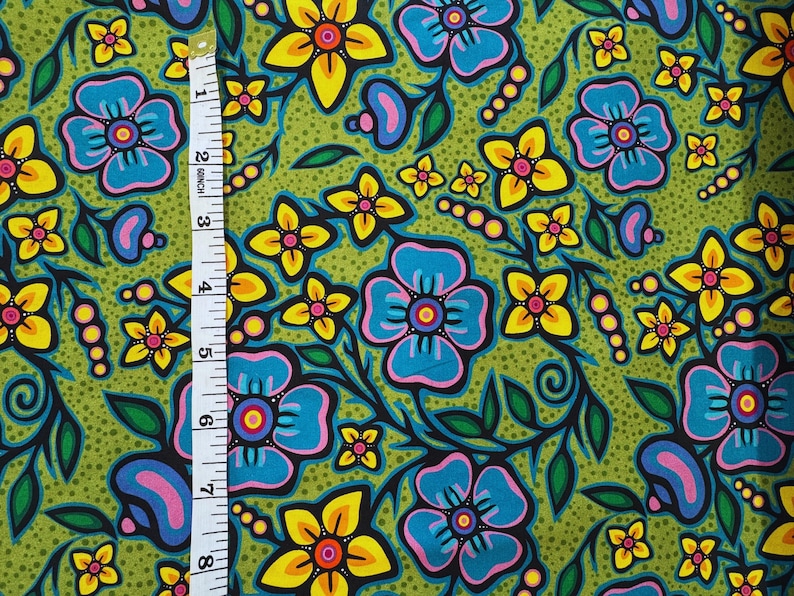 May include: A vibrant fabric with a floral pattern. The design features large blue and yellow flowers with black outlines and pink centers, set against a green dotted background. A measuring tape shows the fabric's dimensions.