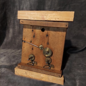 May include: A handcrafted wooden clock with exposed gears and a clock hand. The clock is made from light-coloured wood and has a rectangular form with a base. The gears are metal and are visible on the clock face.