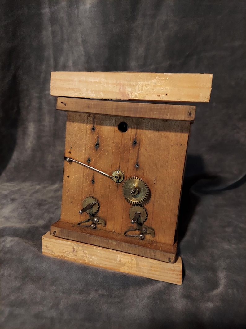 May include: A handcrafted wooden clock with exposed gears and a clock hand. The clock is made from light-coloured wood and has a rectangular form with a base. The gears are metal and are visible on the clock face.