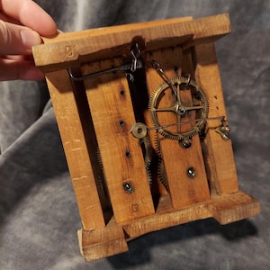 May include: A vintage wooden clock mechanism with visible gears and a pendulum hook. The clock's wooden frame is constructed with vertical and horizontal wooden slats. The gears are made of brass. The clock is held by a hand.