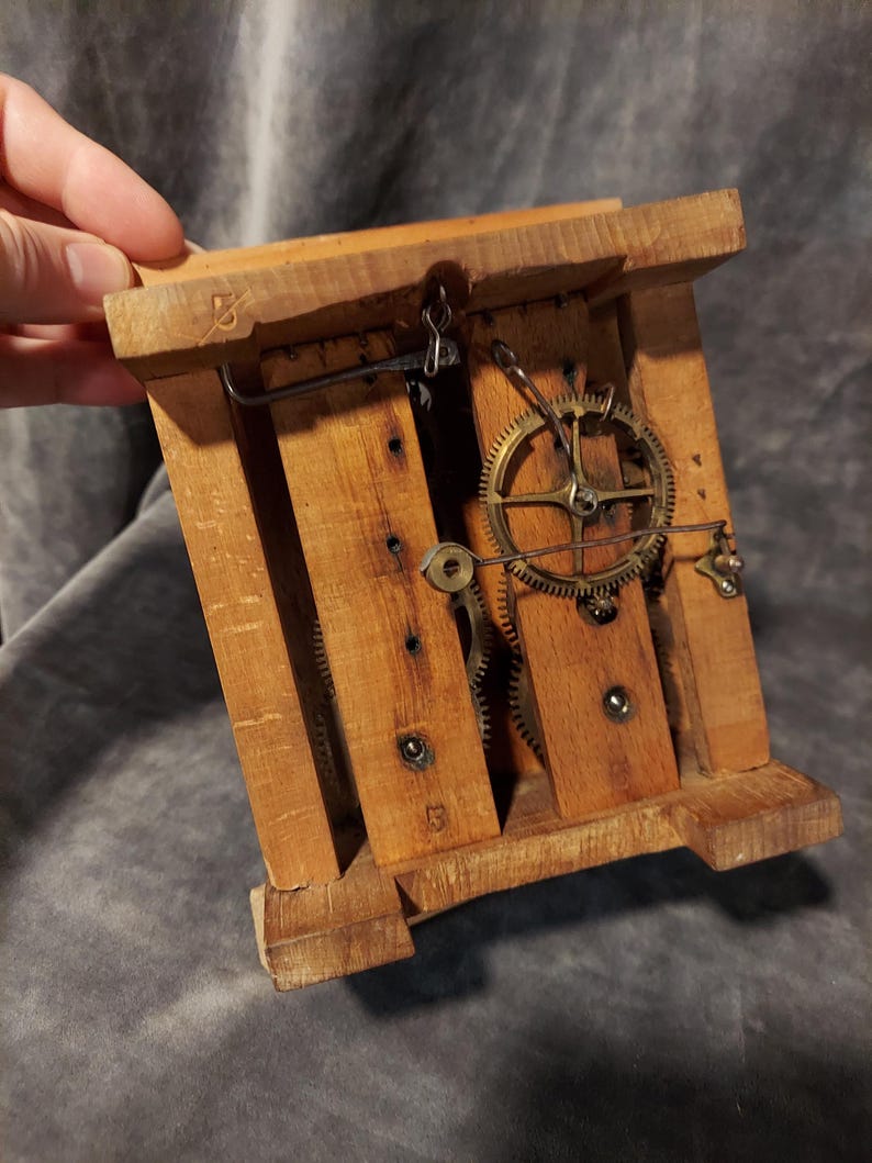 May include: A vintage wooden clock mechanism with visible gears and a pendulum hook. The clock's wooden frame is constructed with vertical and horizontal wooden slats. The gears are made of brass. The clock is held by a hand.