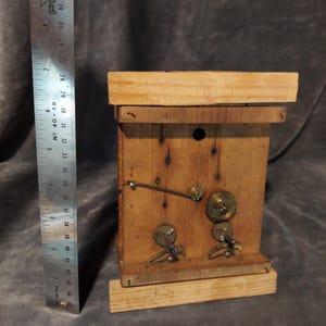 May include: A handmade wooden clock with visible gears and a pendulum. The clock is constructed from light-colored wood and features metal gears and a ruler for scale. The clock's dimensions are approximately 13 cm in height.