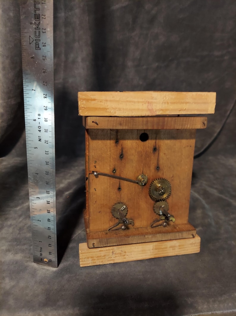 May include: A handmade wooden clock with visible gears and a pendulum. The clock is constructed from light-colored wood and features metal gears and a ruler for scale. The clock's dimensions are approximately 13 cm in height.