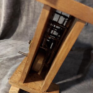 May include: A wooden clock mechanism with exposed gears and internal workings. The clock's wooden frame is angled, displaying the intricate brass gears and components. The clock is made of light brown wood.