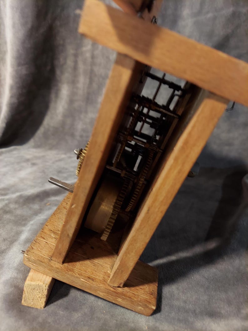 May include: A wooden clock mechanism with exposed gears and internal workings. The clock's wooden frame is angled, displaying the intricate brass gears and components. The clock is made of light brown wood.