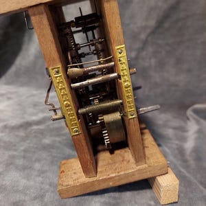 May include: A vintage clock mechanism with visible gears and brass detailing, set within a wooden frame. The intricate internal components of the clock are on display. The wooden frame is a light brown colour.