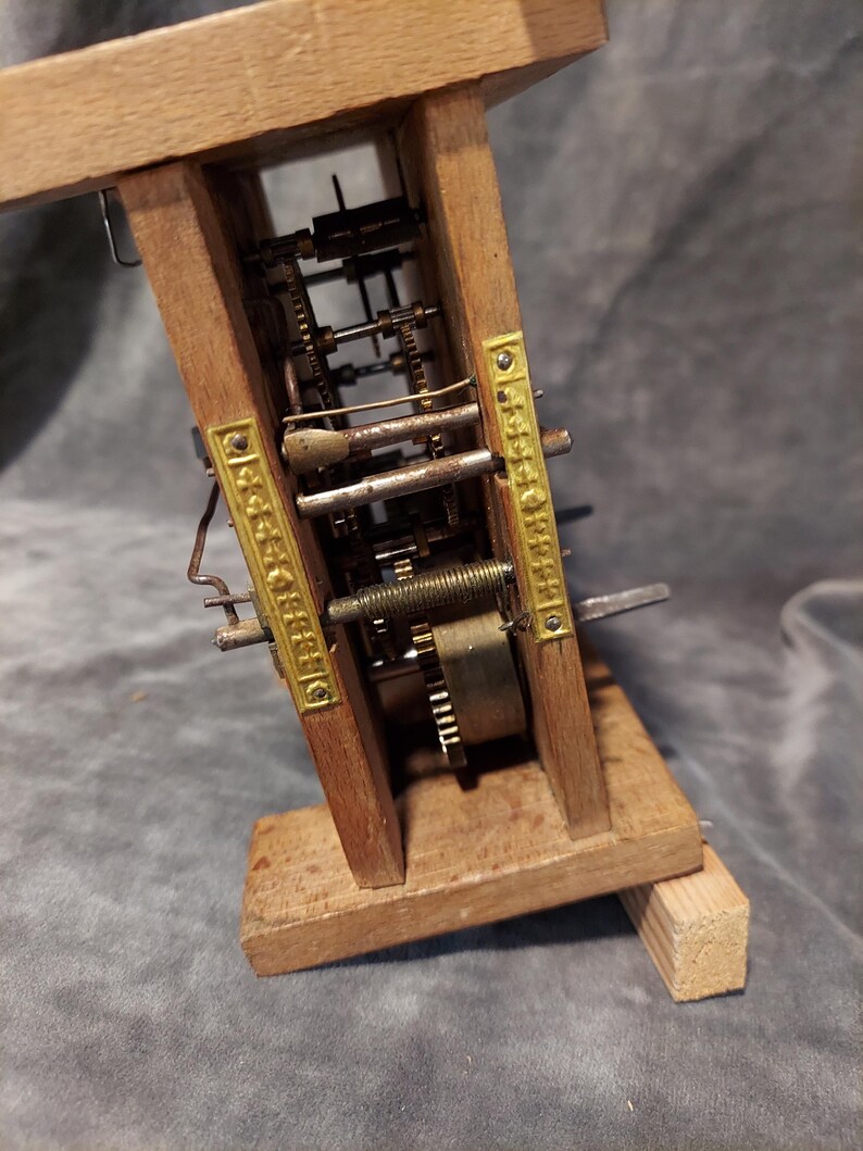 May include: A vintage clock mechanism with visible gears and brass detailing, set within a wooden frame. The intricate internal components of the clock are on display. The wooden frame is a light brown colour.