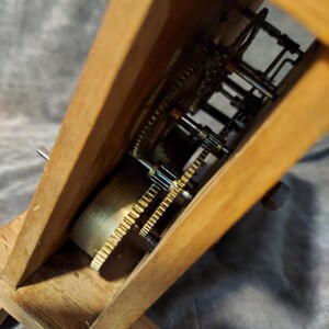 May include: Close-up of a wooden clock mechanism. The clock's internal gears and components are visible within the wooden frame. The gears are made of brass and other metals. The clock is set on a grey surface.