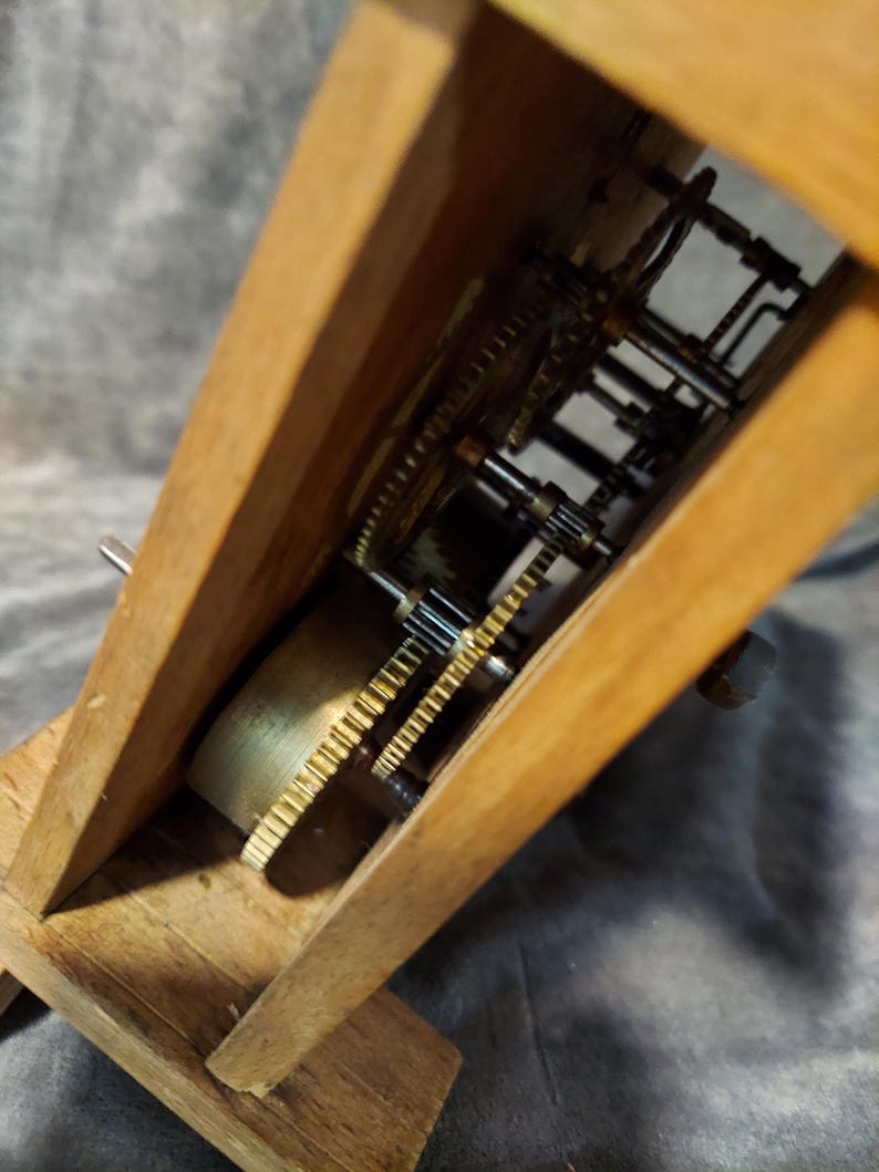 May include: Close-up of a wooden clock mechanism. The clock's internal gears and components are visible within the wooden frame. The gears are made of brass and other metals. The clock is set on a grey surface.