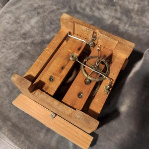 May include: A wooden clock mechanism with exposed gears and a small metal pendulum. The clock's frame is constructed from light-coloured wood, with visible screws and metal components. The gears are brass-coloured, and the overall design is rustic.