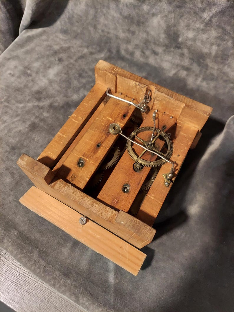 May include: A wooden clock mechanism with exposed gears and a small metal pendulum. The clock's frame is constructed from light-coloured wood, with visible screws and metal components. The gears are brass-coloured, and the overall design is rustic.
