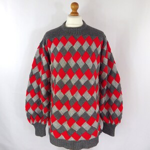 May include: A knitted jumper featuring a diamond pattern in red, grey, and white. It has a high, ribbed collar and cuffs, with long, voluminous sleeves. The garment is displayed on a wooden mannequin. Measurements are in centimeters.
