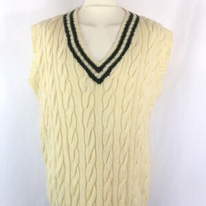 May include: Cream-colored cable knit sweater vest with a deep V-neck and black stripes along the neckline and hem. The sleeveless vest is displayed on a mannequin. The cable knit pattern adds texture to the garment.