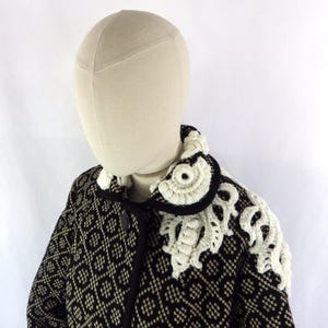 May include: A black and cream patterned coat with a decorative white crocheted collar and shoulder embellishments. The coat features a diamond pattern and black trim, fastened with black buttons. The mannequin head is beige.