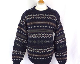 May include: A navy blue patterned sweater with a crew neck. The design includes geometric and floral motifs in brown, grey, and white. The long sleeves have ribbed cuffs. The sweater is displayed on a wooden mannequin.