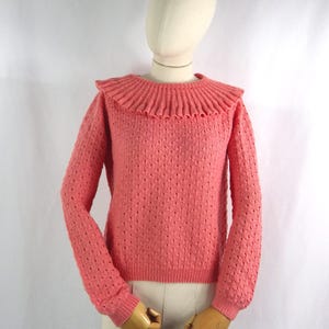 Könnte beinhalten: Ein korallenfarbener Pullover mit einem strukturierten Strickmuster und einem gerüschten Kragen. Das langärmlige Oberteil wird auf einer Schaufensterpuppe präsentiert. Das Design des Pullovers umfasst ein strukturiertes Strickmuster an Ärmeln und Körper.