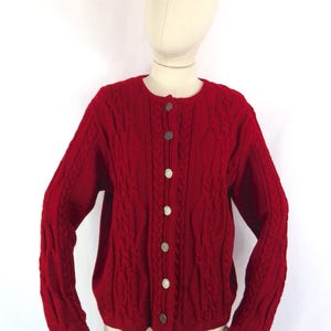 May include: A red cable-knit cardigan with a round neck and long sleeves. The sweater features a button-up front with silver buttons. The garment is displayed on a mannequin.