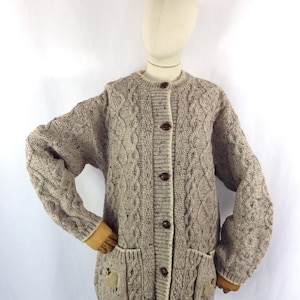 以下が含まれることがあります： 丸いネックラインとボタン留めのベージュのケーブルニットカーディガン。セーターには、羊のアップリケが付いた2つのフロントポケットが付いています。長袖カーディガンはマネキンに表示されています。
