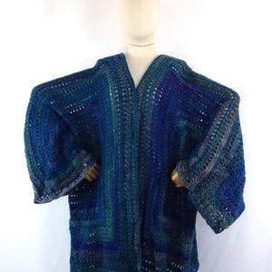 May include: A handmade, open-front crochet cardigan in shades of blue and green. The long-sleeved garment features a V-neck and a loose, flowing design. The crochet stitch creates a textured appearance, and the color transitions add visual interest.
