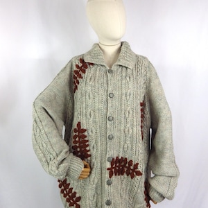 Op de afbeelding: Een grijze kabelgebreide cardigan met bruine geborduurde bladeren. De cardigan heeft een knoopsluiting aan de voorkant.