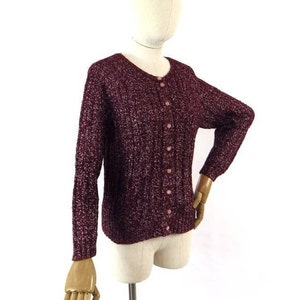 May include: A maroon cardigan with a sparkly texture and pink buttons. The cardigan has long sleeves and a round neckline.