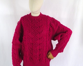 May include: A pink knitted sweater with a textured cable knit pattern and bobbles. The sweater has a crew neck and long sleeves.