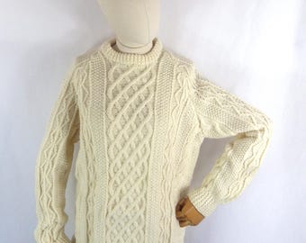 May include: Cream-colored cable knit sweater with a crew neck. The sweater features a central cable design and textured patterns on the sleeves. The garment is displayed on a mannequin against a white background.