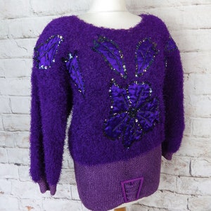 May include: A purple fuzzy sweater with a large flower design made of sequins and satin. The sweater has long sleeves and a ribbed hem.
