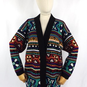 May include: A long, open-front cardigan with a black collar and trim. The sweater has a colourful geometric pattern in teal, red, white, and gold. The garment is displayed on a mannequin.