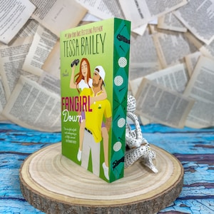 Op de afbeelding: Een boek getiteld "Fangirl Down" van Tessa Bailey. De omslag toont een stel, een vrouw met een verrekijker en een man in een golfshirt. De rug is groen met golf-thema illustraties. Het boek ligt op een houten schijf.